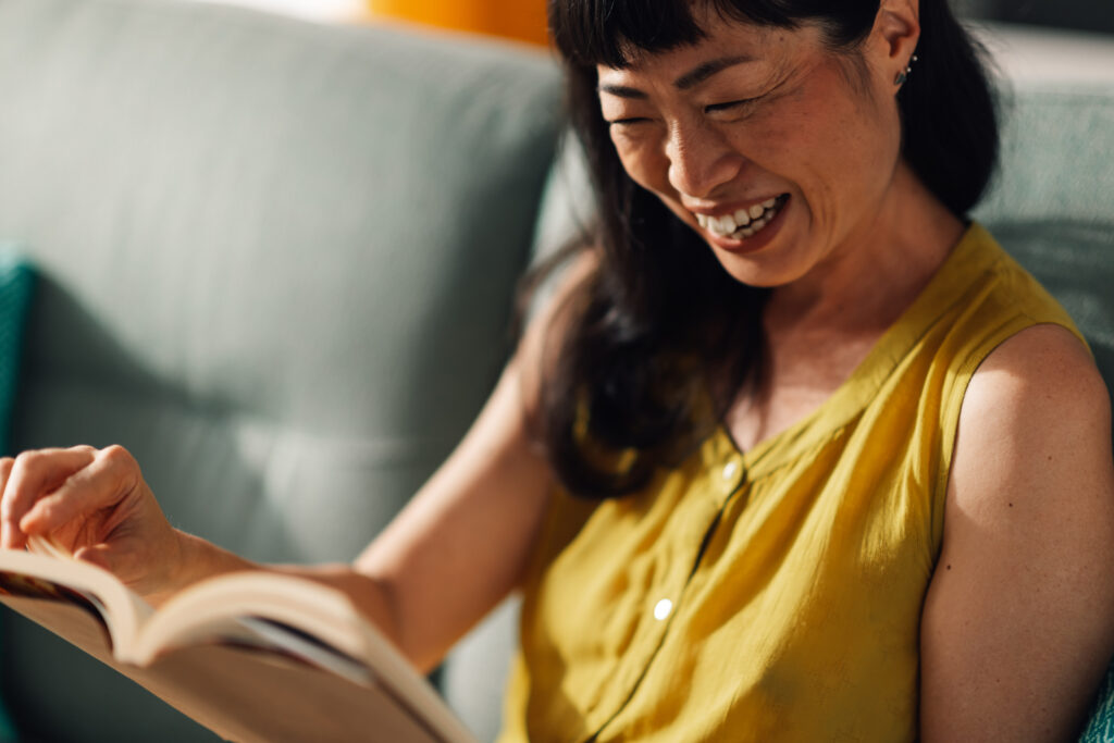 소파에 앉아 환하게 웃으면서 책을 읽는 아시아 여성 │ 편안한 독서와 홈 라이프스타일 이미지